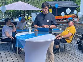 Ein Mann in einem dunklen T-Shirt hält eine kurze Ansprache an einem Stehtisch. Um ihn herum sitzen Gäste auf der Terrasse und hören ihm zu.