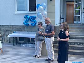 Drei Personen, zwei Frauen und ein Mann, stehen im Gespräch zusammen vor einer Hauswand. Im Hintergrund schmücken blaue und weiße Luftballons sowie eine große silberne Zahl "25" die Fassade.