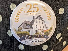 Eine Nahaufnahme zeigt einen runden Ansteck-Button zum Jubiläum. Er trägt die goldene Aufschrift "25 Jahre" und den Schriftzug "Jubiläum Wohnstätte Hotopstraße" über einer Zeichnung des Gebäudes.