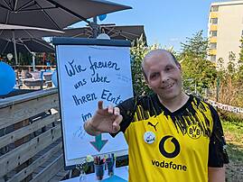 Ein Mann in einem gelb-schwarzen Fußballtrikot lächelt in die Kamera, während er neben einem Flipchart steht. Er macht eine einladende Geste in Richtung eines Gästebuchs.
