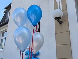 Mehrere hellblaue, blaue und weiße Luftballons sind an der Fassade eines Hauses befestigt. Darunter ist eine große, blaue Folienzahl "25" zu sehen, die auf das Jubiläum hinweist.