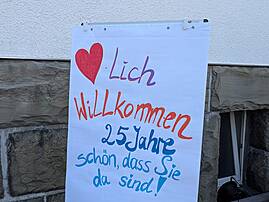 Ein handgemaltes Plakat auf einer Staffelei begrüßt die Gäste. Darauf steht in bunten Buchstaben: "Herzlich Willkommen 25 Jahre schön, dass Sie da sind!".