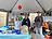 Bild 1: Unter einem Pavillon stehen drei Personen am Stand der Lebenshilfe vor einem Tisch mit Informationsmaterial, Süßigkeiten und einem bunten Glücksrad. Im Hintergrund sind Luftballons zu sehen.