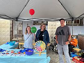 Bild 1: Unter einem Pavillon stehen drei Personen am Stand der Lebenshilfe vor einem Tisch mit Informationsmaterial, Süßigkeiten und einem bunten Glücksrad. Im Hintergrund sind Luftballons zu sehen.