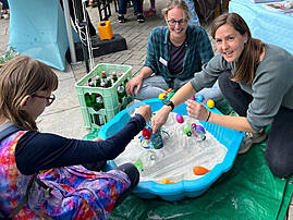Bild 2: Eine junge Person spielt gemeinsam mit zwei Mitarbeitenden an einer blauen Wanne mit Sand und bunten Gegenständen. Die Szene zeigt eine aktive Begegnung am Stand der Lebenshilfe beim FunFestival.