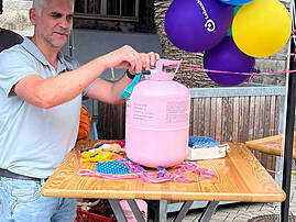 Bild 3: Eine Person befestigt bunte Luftballons an einem Tisch neben einem großen rosafarbenen Ballonbehälter. Der Stand ist mit weiteren Materialien und Lebenshilfe-Luftballons geschmückt.