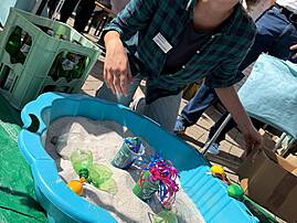 Bild 4: Eine lächelnde Mitarbeiterin der Lebenshilfe sitzt neben einer blauen Sandwanne und blickt in die Kamera. Im Hintergrund sind der Pavillon, eine Getränkekiste und weitere Besucher*innen des Festivals zu sehen.