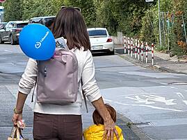 Bild 5: Eine erwachsene Person geht mit einem Kind über eine Straße und trägt einen Rucksack mit einem blauen Lebenshilfe-Luftballon. Im Hintergrund sind parkende Autos, Bäume und ein Parkplatz für Menschen mit Behinderung zu sehen.