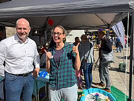 Bild 6: Zwei lächelnde Mitarbeitende stehen am Lebenshilfe-Stand, während eine Mitarbeiterin beide Daumen nach oben zeigt. Im Hintergrund sind Besucher*innen, Lebenshilfe-Luftballons, Informationsmaterial und eine blaue Sandwanne zu sehen.