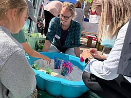 Bild 7: Eine Mitarbeiterin sitzt gemeinsam mit zwei weiteren Personen an einer blauen Sandwanne mit bunten Spielmaterialien unter einem Pavillon.