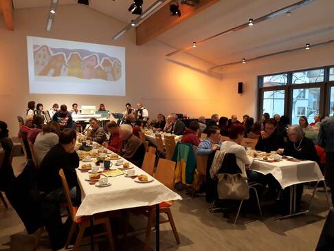Viele Menschen sitzen in einem Raum an gedeckten Tischen.