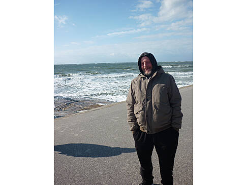 Ein Mann steht mit einer dicken Jacke und Händen in der Hosentasche am Strand, er hat eine Kapuze an.