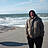 Ein Mann steht mit einer dicken Jacke und Händen in der Hosentasche am Strand, er hat eine Kapuze an.