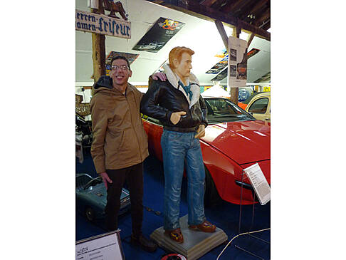Ein Mann steht neben einer lebensgroßen Figur und lacht. Er hat seine Hand auf der Schulter. Sie Stehen neben einem roten Auto.