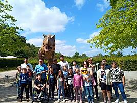 Eine große Gruppe von Kindern, Jugendlichen und Erwachsenen posiert für ein Gruppenfoto im Freien. Hinter ihnen steht ein lebensgroßes Modell eines Tyrannosaurus Rex mit geöffnetem Maul.