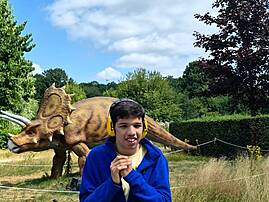 Ein Jugendlicher in einer blauen Jacke steht lächelnd vor dem Modell eines Triceratops in einer Parklandschaft. Er trägt gelbe Kopfhörer um seinen Hals.