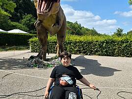 Eine junge Person sitzt lächelnd in einem Rollstuhl vor der großen Nachbildung eines Tyrannosaurus Rex. Die Dinosaurierfigur hat ihr Maul weit aufgerissen und scheint zu brüllen.