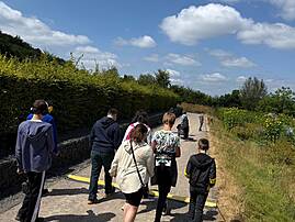 Eine Gruppe von Menschen spaziert von hinten gesehen auf einem Weg durch eine Parkanlage. Links und rechts des Weges wachsen grüne Hecken und Gräser unter einem blauen Himmel.