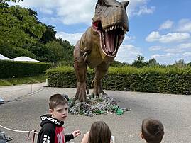 Drei Kinder stehen vor dem riesigen Modell eines Tyrannosaurus Rex und betrachten es. Der Dinosaurier ist sehr detailreich dargestellt und hat sein Maul weit geöffnet.