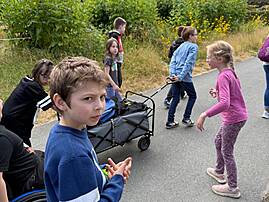 Mehrere Kinder und Jugendliche unterhalten sich auf einem Weg in einem Park, neben ihnen steht ein Bollerwagen. Ein Junge im Vordergrund blickt direkt in die Kamera.