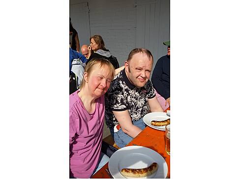 Zwei Personen haben eine Wust auf dem Teller und sitzen in der Sonne.