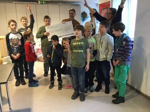 Mehrere Kinder stehen mit drei Erwachsenen in einem Raum und haben einen Scheck.