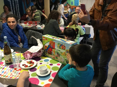 Mehrere Personen sitzen an Tischen, Kinder packen Geschenke aus.