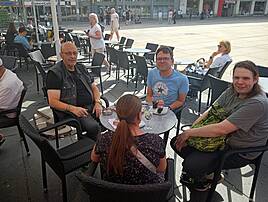 Vier Personen sitzen an einem Tisch im Freien, trinken etwas und führen ein Gespräch in entspannter Atmosphäre. Im Hintergrund sind weitere Gäste und ein belebter Platz zu sehen