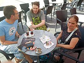 Drei Menschen mit und ohne Unterstützungsbedarf sitzen an einem Tisch in einer Eisdiele, genießen Getränke und unterhalten sich.