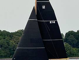 Ein klassisches Segelboot mit zwei großen schwarzen Segeln, beschriftet mit „K-14", fährt auf ruhigem Wasser vor einer bewaldeten Küste.