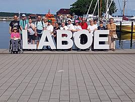 Eine Gruppe von etwa 15 Personen steht hinter großen weißen Buchstaben, die das Wort „LABOE" bilden, am sonnigen Hafen von Laboe.