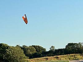 Ein Kitesurfer gleitet nahe am Ufer über Wellen, während ein orangefarbener Drachen hoch in einem blauen Sommerhimmel steht.