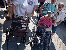 Eine Gruppe Menschen am Hafen, darunter eine Person im Rollstuhl und eine mit Rollator, wartet in der Sonne auf einem gepflasterten Weg.