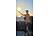 in junger Mann in orangefarbenem T-Shirt steht bei Sonnenuntergang am Strand und streckt eine Hand aus, während ein Kitesurfer-Drachen am Abendhimmel zu sehen ist.