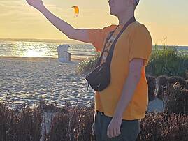in junger Mann in orangefarbenem T-Shirt steht bei Sonnenuntergang am Strand und streckt eine Hand aus, während ein Kitesurfer-Drachen am Abendhimmel zu sehen ist.