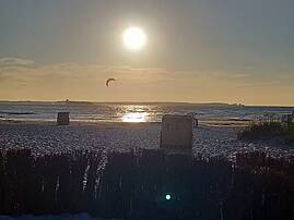 Abendlicher Sonnenuntergang an einem Sandstrand mit Strandkörben und einem Kitesurfer-Drachen am Horizont, aufgenommen durch Schilfgras.