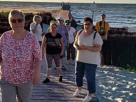 Mehrere Personen spazieren in einer Reihe über einen Holzsteg am Strand, im Hintergrund ist das Meer zu sehen.