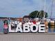 Eine Gruppe von etwa 15 Personen steht hinter großen weißen Buchstaben, die das Wort „LABOE" bilden, am sonnigen Hafen von Laboe.