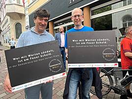 Jörg Bachmann (links) und Sven Sieburg (rechts) stehen auf einer Straße und halten schwarze Schilder hoch. Auf den Schildern steht der Slogan "Wer mit Worten zündelt, ist am Feuer Schuld. In der Demokratie hat Hass keinen Platz."