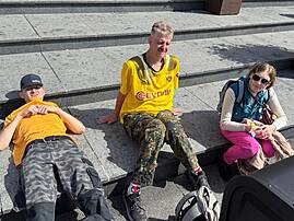 Bild 1: Drei Personen sitzen beziehungsweise liegen auf breiten Steinstufen vor einem Gebäude mit großen Blumenkübeln.