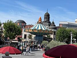 Bild 4: Auf einem sonnigen Platz steht ein blau-weiß gestreiftes Karussell vor mehreren Kuppelgebäuden. Im Vordergrund spenden rote Sonnenschirme Schatten, während Menschen über den Platz gehen.