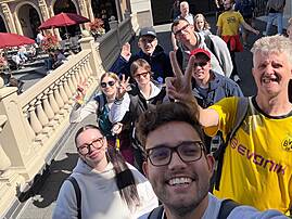 Bild 6: Eine Gruppe von sieben Personen macht auf einer Außentreppe ein Selfie und zeigt teilweise Friedenszeichen. Im Hintergrund sind ein Gebäude, rote Sonnenschirme und weitere Gäste zu sehen.