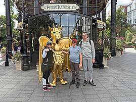 Bild 8: Drei Personen stehen vor einer goldfarbenen Drachenfigur am Eingang eines Fahrgeschäfts. Über dem Eingang steht „Pferdekarussell“.