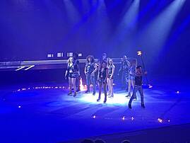 Bild 11: Mehrere Performer*innen stehen auf einer großen Bühne in blauem Licht, umgeben von kleinen Flammen und dem Publikum.