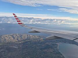 Blick aus dem Flugzeug auf Mallorca mit Küste, Bergen und Wolken. Im Vordergrund ist der Flugzeugflügel zu sehen.