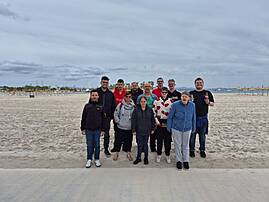 Eine Reisegruppe steht gemeinsam an einem Strand auf Mallorca. Der Himmel ist bewölkt, im Hintergrund sind Meer und Strandliegen zu sehen.