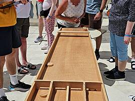 Mehrere Personen spielen gemeinsam ein Holzspiel im Freien. Die Gruppe steht konzentriert um das Spielbrett auf einer sonnigen Terrasse.