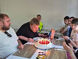 Eine Gruppe feiert Geburtstag an einem Tisch. Eine Person pustet Kerzen auf einer Torte aus, während die anderen zusehen.
