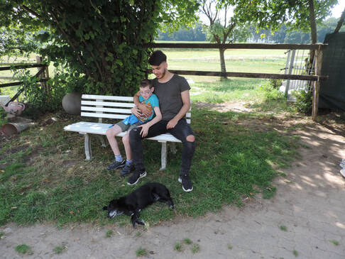 Ein Mann hält einen Jungen im Arm, sie sitzen auf einer weißen Bank unter einem Baum im Schatten, davor liegt ein dunkler Hund.