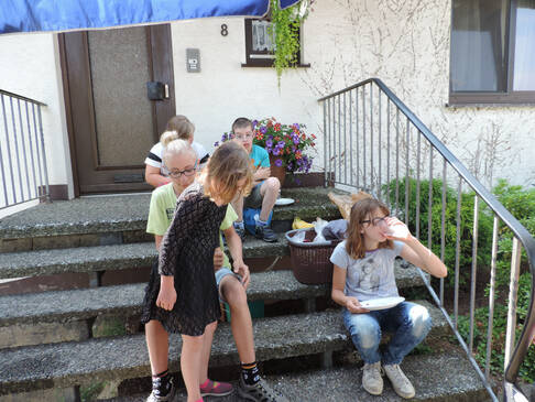 Personen sitzen vor einem Haus, im Schatten auf einer Treppe.